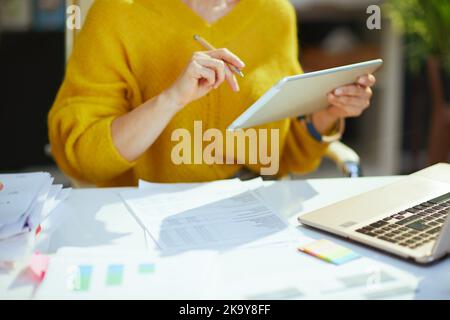Nahaufnahme der Inhaberin eines kleinen Unternehmens mittleren Alters in gelbem Pullover mit Tablet-PC und der Arbeit mit Dokumenten im grünen Büro. Stockfoto