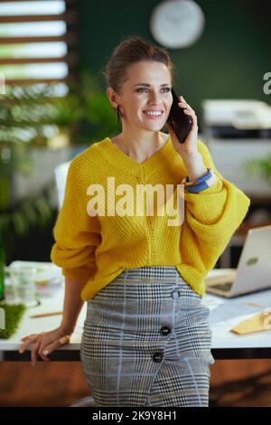 Glückliche elegante 40 Jahre alte Kleinunternehmerin Frau in gelbem Pullover im modernen grünen Büro im Gespräch auf einem Smartphone. Stockfoto