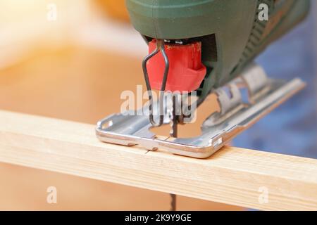 Die Hände eines Zimmermanns mit einer elektrischen Stichsäge in den Händen, die ein Stück Holz abschneidet. Männliche Hände mit Laubsäge zum Schneiden von Holz. Ein Zimmermann schneidet Holz mit einer Stichsäge in einer heimischen Werkstatt. Stockfoto