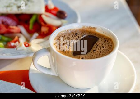 Griechischer Kaffee auf dem Esstisch mit buntem griechischem Salat, der im Hintergrund verschwommen ist Stockfoto