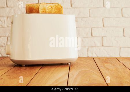 Weißer elektrischer Toaster und fertig gebackene Brotscheiben auf dem hölzernen Küchentisch. Kochen und Haushaltswaren Technologien Hintergründe Stockfoto