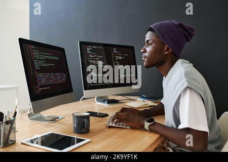 Junger seriöser afroamerikanischer männlicher Programmierer, der über neue Software vor Computerbildschirmen mit kodierten Daten im Büro arbeitet Stockfoto