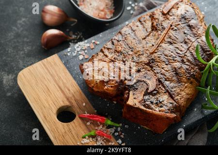 Rohes Steak, T-Bone oder Porterhouse. Steak-T-Bone oder Porterhouse in Sauce mit Gewürzen, Salz und Rosmarin auf einem schwarzen Keramikplatte auf Betonboden. Stockfoto