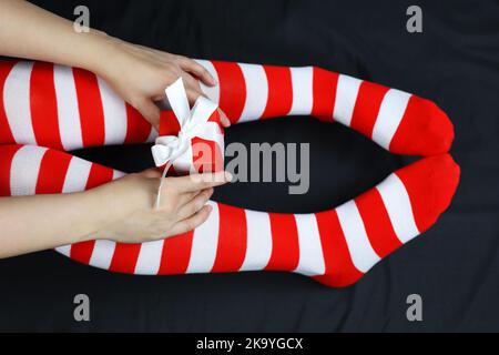 Mädchen in Weihnachtskniesocken sitzend mit roter Geschenkbox auf schwarzem Bettzeug. Weibliches Outfit zur Neujahrsfeier Stockfoto