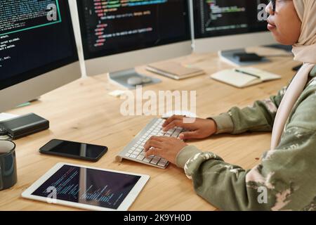 Junge muslimische Praktikantin in Hijab tippen auf der Tastatur, während Daten auf Computerbildschirmen am Arbeitsplatz mit Handy und Tablet decodieren Stockfoto