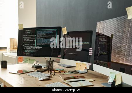 Gruppe von Monitoren mit kodierten Daten und Haftnotizpapieren auf Bildschirmen, die im Büro moderner IT-Ingenieure auf einem Holztisch stehen Stockfoto