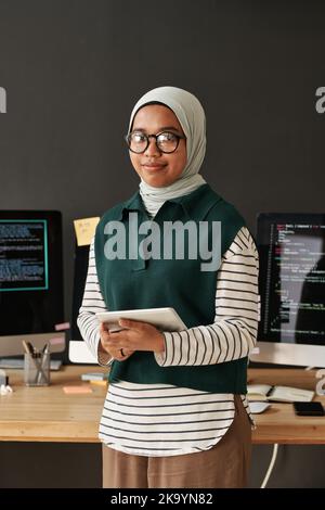 Junge erfolgreiche muslimische Geschäftsfrau oder Praktikantin mit Tablet in den Händen, die Kamera anschaut, während sie am Arbeitsplatz mit Computern steht Stockfoto