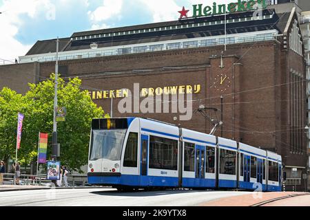 Amsterdam, Niederlande - August 2022: Moderne elektrische Straßenbahn in Amsterdam, die an der alten Heineken-Brauerei vorbeiführt, die heute ein Museum und eine Touristenattraktion ist. Stockfoto