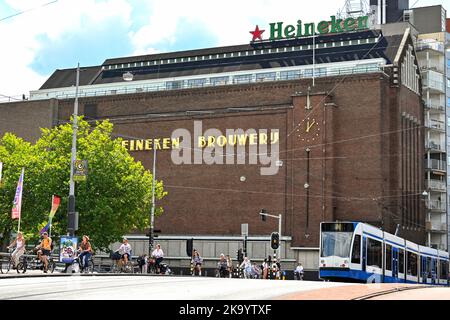 Amsterdam, Niederlande - August 2022: Moderne elektrische Straßenbahn in Amsterdam, die an der alten Heineken-Brauerei vorbeiführt, die heute ein Museum und eine Touristenattraktion ist. Stockfoto