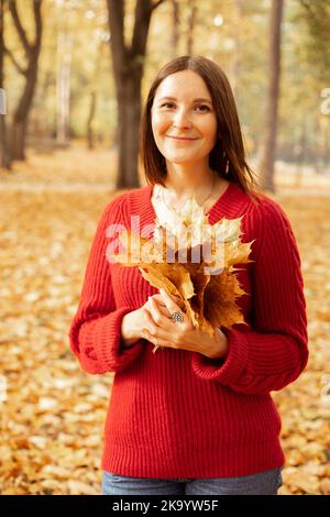 Portrait der fröhlichen hübschen Frau in gemütlichen roten Strickpullover mit Strauß von gelben Blättern isoliert auf goldenen Wald Hintergrund. Glückliche junge Dame Stockfoto