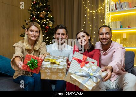 Weihnachtsporträt mit vier Freunden, einer vielfältigen Gruppe von Menschen, die die Kamera betrachten und lächelnd Geschenke für das neue Jahr halten, Gäste zu Hause auf dem Sofa, um in der Nähe des Weihnachtsbaums zu feiern. Stockfoto