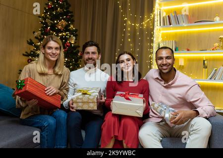 Weihnachtsporträt mit vier Freunden, einer vielfältigen Gruppe von Menschen, die die Kamera betrachten und lächelnd Geschenke für das neue Jahr halten, Gäste zu Hause auf dem Sofa, um in der Nähe des Weihnachtsbaums zu feiern. Stockfoto