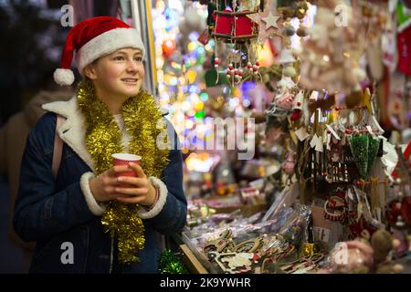 Teenager-Mädchen in Santa hat zu Fuß auf dem Weihnachtsmarkt mit einer Tasse warmen Getränk Stockfoto