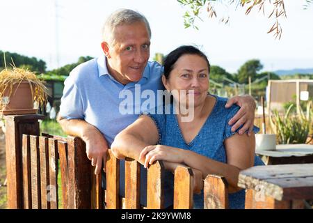 Älterer Mann, der seine Frau umarmt, während er in der Nähe des Holzzauns auf dem Gehöft steht Stockfoto