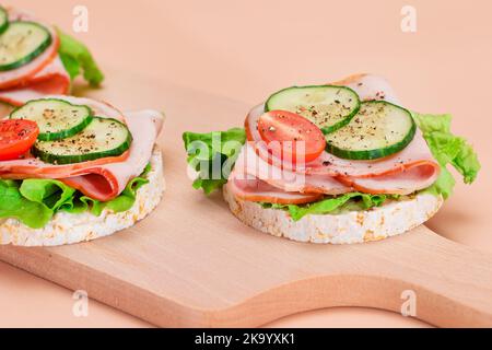 Leichtes Frühstück. Schnelle und gesunde Sandwiches. Reiskuchen mit Schinken, Tomaten, frischem Gurken und grünem Salat auf Holzschneidebron. Beigefarbener Hintergrund Stockfoto