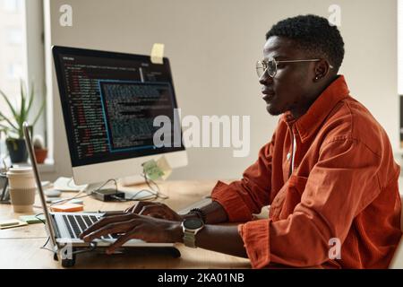 Seitenansicht eines jungen schwarzen Mannes in eleganter Freizeitkleidung und einer Brille, die Daten entschlüsselt, während er vor dem Laptop am Arbeitsplatz sitzt Stockfoto