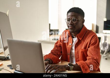 Junger afroamerikanischer IT-Ingenieur oder Webdesigner, der beim Entschlüsseln von Informationen oder Erstellen einer neuen Website auf den Laptop-Bildschirm schaut Stockfoto