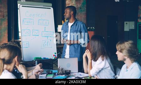 Der afroamerikanische Business Trainer arbeitet mit einem Whiteboard, das auf das Diagramm zeigt und spricht, während die Mitarbeiter des Unternehmens lernen, lernen und sprechen, während sie am Tisch im Büro sitzen. Stockfoto