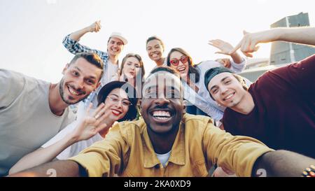 Aus der Sicht des hübschen afroamerikanischen Mannes, der mit seinen glücklichen Freunden Selfie macht, blicken die Leute auf die Kamera, lächeln und posieren mit Flaschen auf einer lustigen Dachparty. Stockfoto