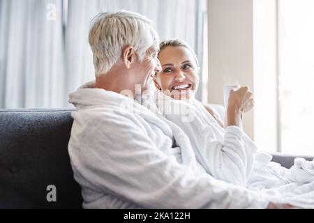 Wir verdienen es, ab und zu verwöhnt zu werden. Ein fröhliches Paar mittleren Alters, das sich zusammen entspannt, während es Bademäntel trägt und auf einer Couch im Spa sitzt Stockfoto