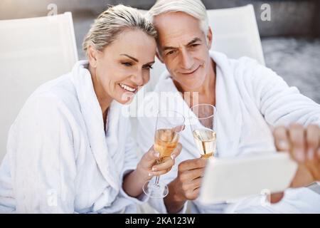 Ein fröhliches Paar mittleren Alters, das ein Selbstporträt zusammen, während Sie ein Glas Champagner draußen in einem Spa genießen während der Stockfoto