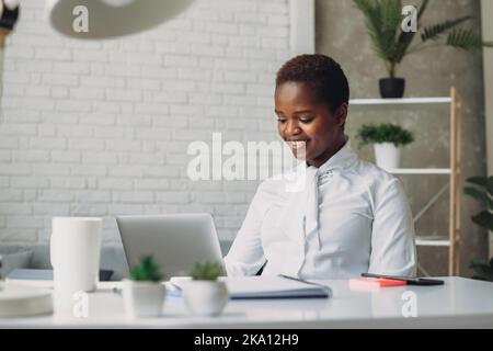Fokussierte junge afroamerikanische Geschäftsfrau, die im modernen Büro eines Unternehmens auf den Laptop-Bildschirm schaut und auf die Computertastatur tippt. Kommunikation Stockfoto