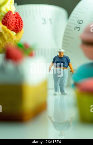 Miniaturfigur eines übergewichtigen Mannes gegen Maßband und ungesunde Lebensmittel. Stockfoto