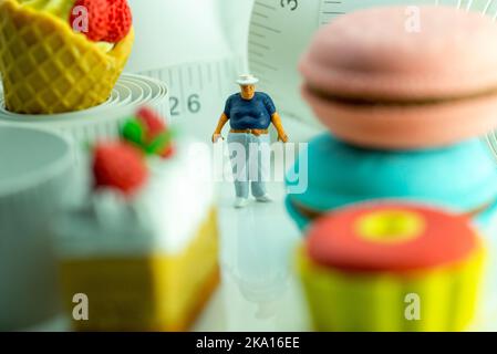 Miniaturfigur eines übergewichtigen Mannes gegen Maßband und ungesunde Lebensmittel. Stockfoto