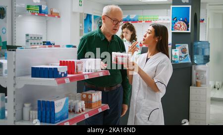 Medizinisches Personal, das dem älteren Mann kardiologische Medikamente erklärt und im Apothekengeschäft über Gesundheitsmedizin spricht. Frau, die Klientin bei der Behandlung und Pharmazie, medizinische Hilfe hilft. Stockfoto