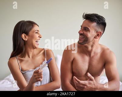 Sie sind begeistert von den guten Nachrichten. Ein junges Ehepaar begeistert sich über die Ergebnisse eines Schwangerschaftstests in ihrem Schlafzimmer zu Hause. Stockfoto