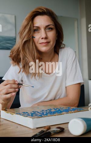 Nahaufnahme Porträt einer glücklichen kreativen jungen Frau, die lächelt und direkt auf die Kamera schaut, während sie in ihrem Atelier auf Leinwand malte. Person, die hält Stockfoto