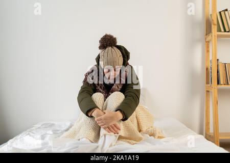 Eine wütende junge Frau, die warm in einem kalten Haus gekleidet ist, sitzt auf dem Bett und fühlt sich im Haus kalt an Stockfoto