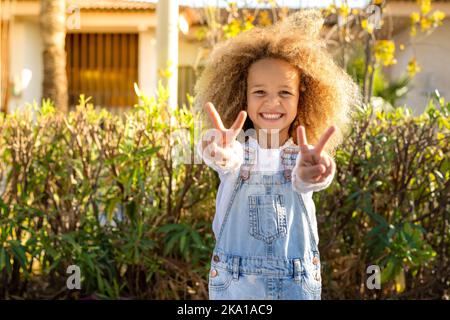 Fröhliches afroamerikanisches Mädchen mit lockigen Haaren, das im Sommerpark zwei Finger zeigt, während es auf die Kamera schaut Stockfoto