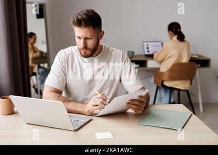 Junger ernsthafter Mann, der sich im Copybook Notizen macht und während des Online-Unterrichts mit dem Tutor auf den Laptop-Bildschirm schaut Stockfoto