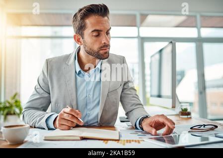 Ein hübscher junger Geschäftsmann, der Notizen schreibt, während er ein digitales Tablet in einem Büro benutzt. Stockfoto