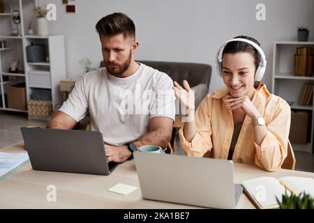 Glückliche junge Frau mit Kopfhörern, die sich im Videochat vor dem Laptop unterhielt, während sie neben einem ernsthaften bärtigen Mann am Schreibtisch saß Stockfoto