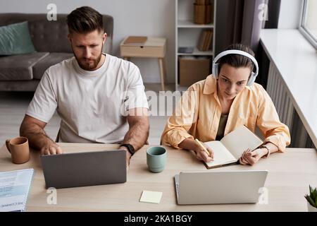 Junge Frau mit Kopfhörern, die sich im Copybook Notizen macht, während sie beim Online-Unterricht neben einem ernsthaften Mann am Schreibtisch vor dem Laptop sitzt Stockfoto