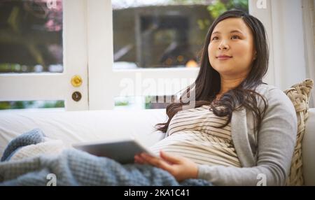 Nicht lange, bis das kleine kommt. Eine Schwangere, die nachdenklich aussieht, während sie zu Hause ein digitales Tablet benutzt. Stockfoto