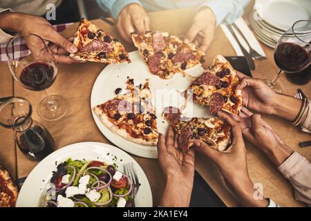 Das Abendessen wird serviert. Eine Gruppe von nicht erkennbaren Personen, die in einem Restaurant sitzen und dabei jeweils ein Stück Pizza greifen, wird in einem hohen Winkel aufgenommen. Stockfoto