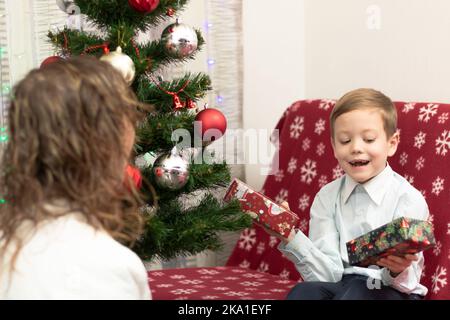 Mutter schenkt ihrem siebenjährigen Sohn zum Neujahr Geschenke in Schachteln vor dem Hintergrund eines Weihnachtsbaums zu Hause. Selektiver Fokus. Hochformat Stockfoto