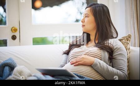 Eine Schwangere, die nachdenklich aussieht, während sie zu Hause ein digitales Tablet benutzt. Stockfoto