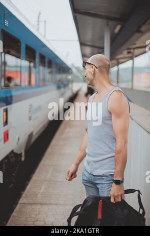 Junger Mann in einem T-Shirt auf dem Bahnsteig, der mit dem Handy auf einen Zug wartet. Mann am Bahnhofsplatz mit Tasche und Mobiltelefon Stockfoto
