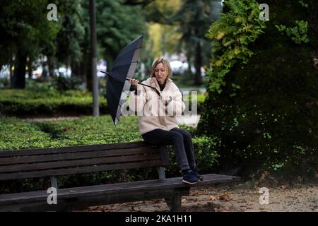 Eine Frau sitzt auf einer Parkbank. Bei Regenwetter brach ein Regenschirm zusammen. Stockfoto