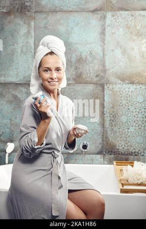 Frau im Spa-Bademantel sitzt auf der Badewanne, halten Gesichtscreme und schrubben in der Hand, lächelnd schaut zur Kamera. Stockfoto