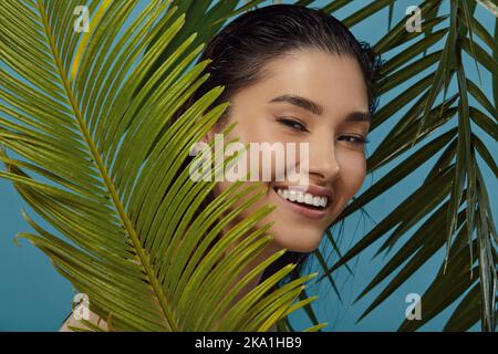 Verspieltes Mädchen guckt hinter Palmblättern hervor und lächelt. Stockfoto