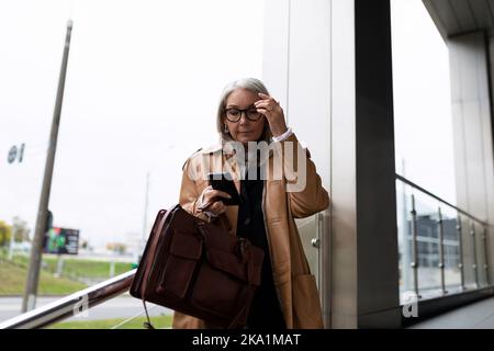 Eine ältere Geschäftsfrau mittleren Alters mit Aktentasche und Telefon in den Händen ist in Eile, sich zu treffen und außerhalb des Bürokonzepts zu arbeiten Stockfoto