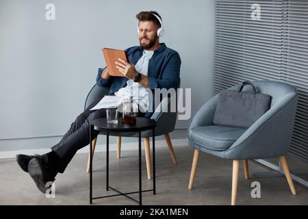 Fröhlicher junger Mann in Kopfhörern und Casualwear mit digitalem Tablet, während er im Sessel sitzt und im Videochat kommuniziert Stockfoto