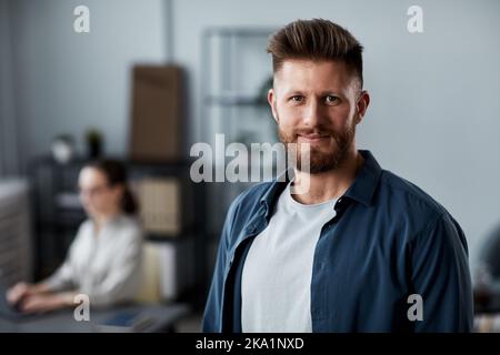 Junger erfolgreicher Chief Executive Officer oder Manager in Casualwear, der die Kamera anschaut, während er im Büro gegen seinen Kollegen steht Stockfoto
