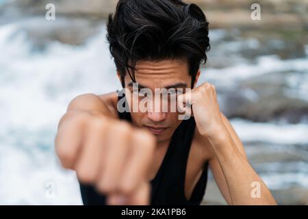 Intensive und fokussierte asiatische 30s Mann üben Kampfkunst und Selbstverteidigung am Meer im Sommer. Konept für Krafttraining Stockfoto