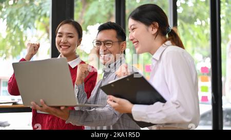 Überglücklich feiert der Erwachsene asiatische Geschäftsmann seinen Projekterfolg mit seinen Teamkollegen im Büro und zeigt triumphale Hände. Teamwork-Konzept Stockfoto